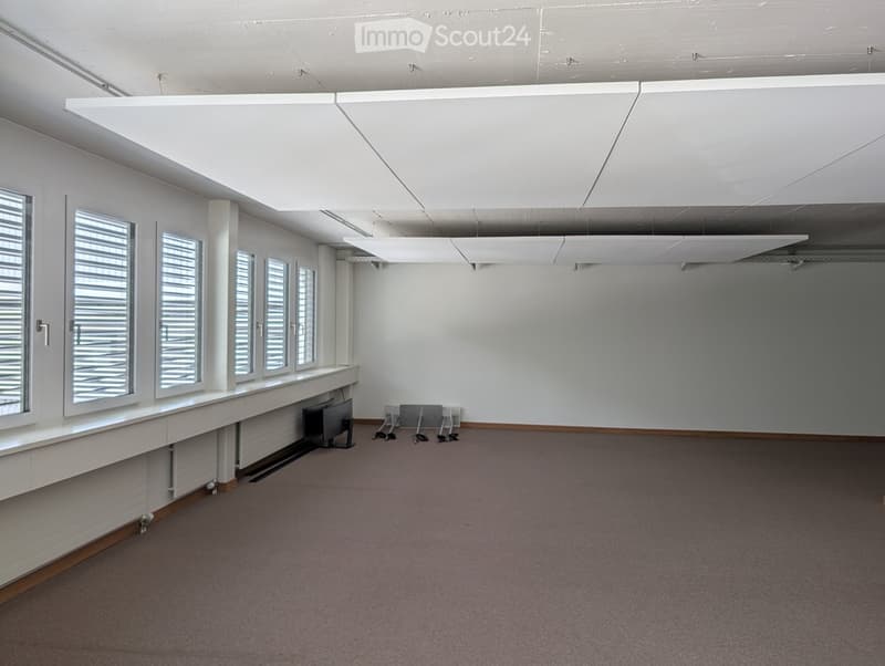 Empty room with white walls, large windows with blinds, grey carpet, and a long white bench along one wall.