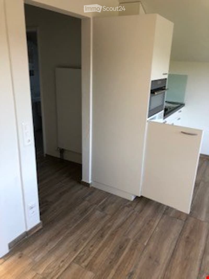 Empty room with wooden flooring, white walls, and a partially visible kitchen area with cabinets and appliances in the background.