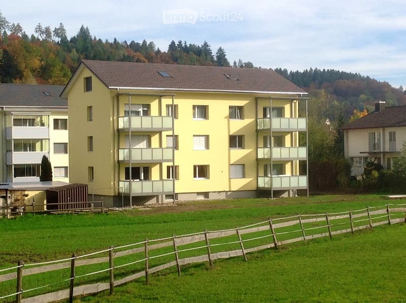 A multi-story yellow apartment building with balconies, surrounded by a wooden fence and green lawn, with trees and hills in the background.