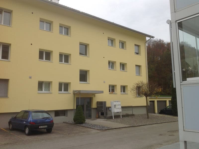 A yellow multi-story building with multiple windows, a covered entrance, and a small parking area with a blue car. There is a tree and a signboard near the entrance.