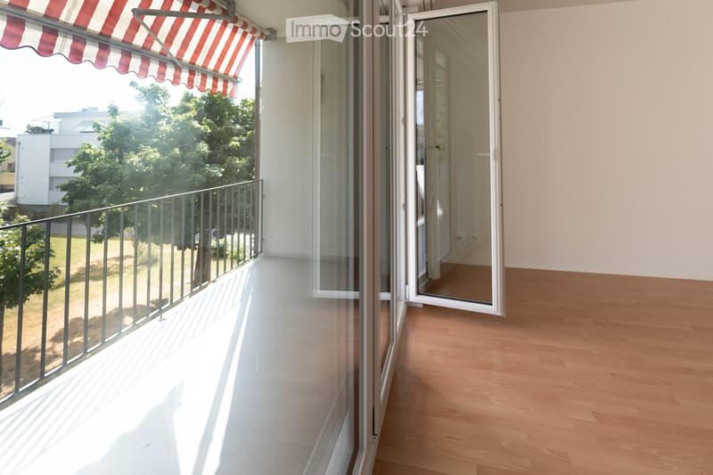 Balcony with wooden floor, glass sliding doors, metal railing, and striped awning. The view shows greenery and a neighboring building.