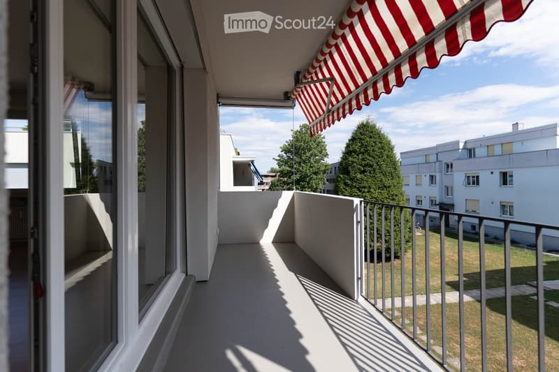 Balcony with a red and white striped awning, metal railing, and view of a green lawn and surrounding buildings.
