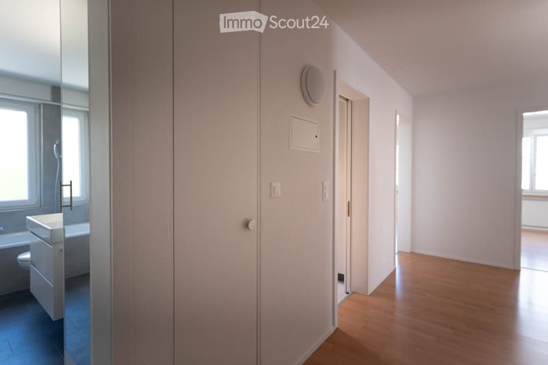 A clean, modern hallway with light wooden flooring, white walls, and several doors leading to different rooms. A small bathroom is visible to the left with a sink, toilet, and bathtub.