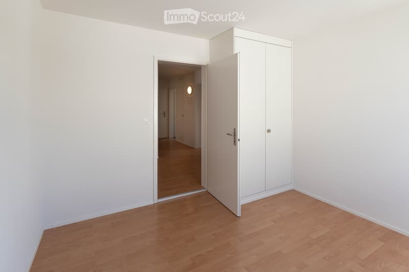 Empty room with light wooden flooring, white walls, and a built-in white wardrobe with mirrored doors. An open doorway leads to a hallway with warm lighting.