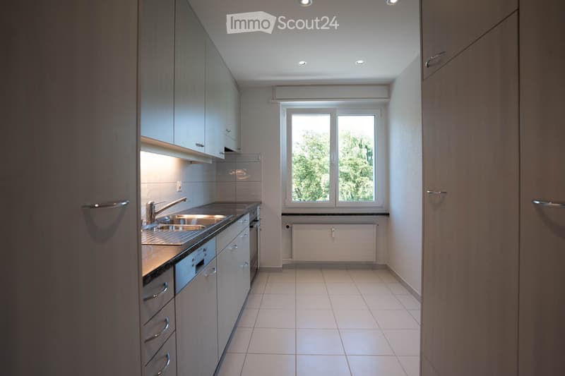 Modern kitchen with light wood cabinets, stainless steel sink, dishwasher, and tiled floor. Large window provides natural light.