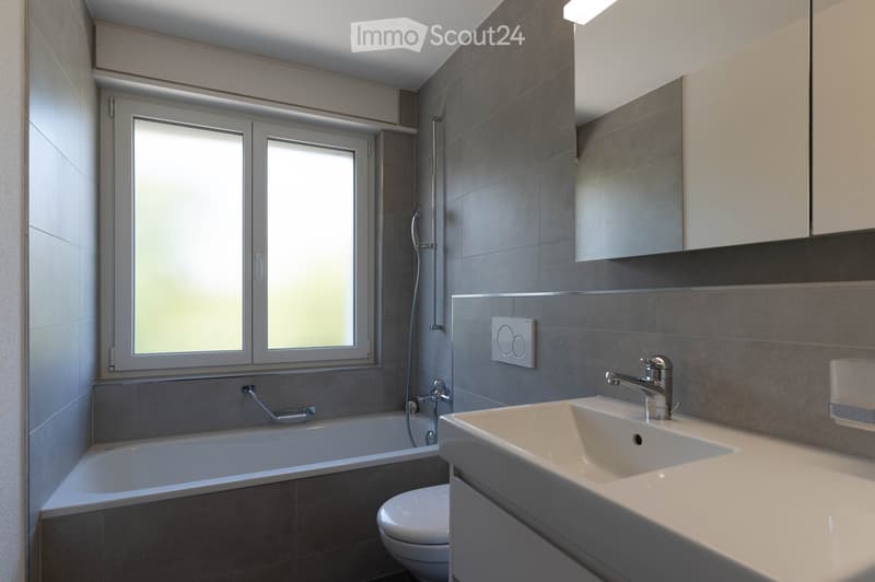 Modern bathroom with a bathtub, sink, toilet, and a large mirror above the sink. The room features grey tiled walls and a window with frosted glass panes.
