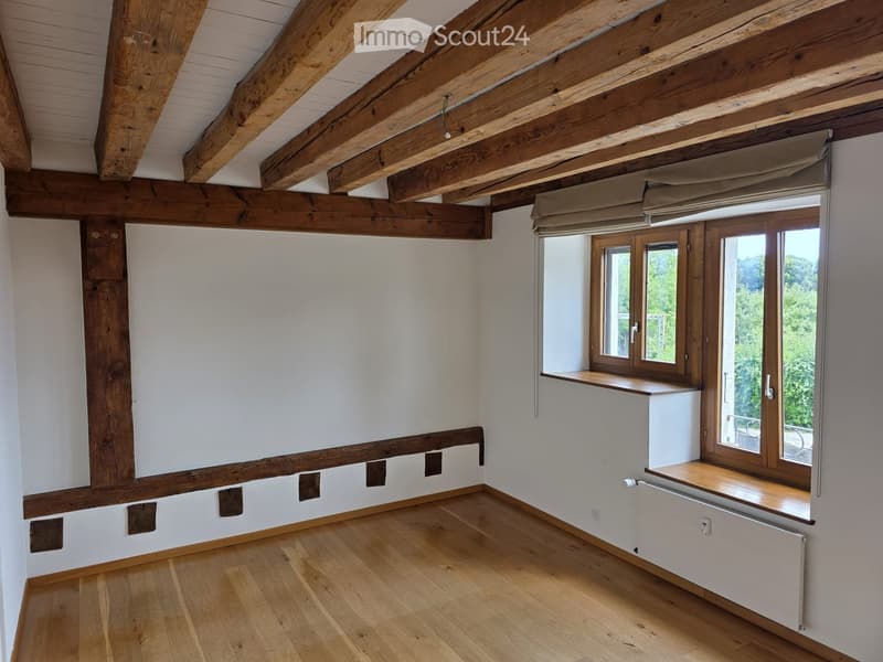 Empty room with wooden beams on the ceiling, wooden floor, large windows with wooden frames, and white walls.