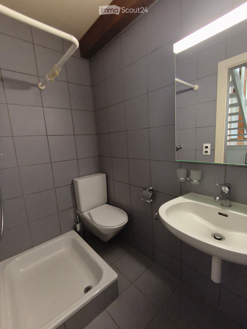 Small bathroom with grey tiled walls, a toilet, a sink, a bathtub, and a mirror above the sink.
