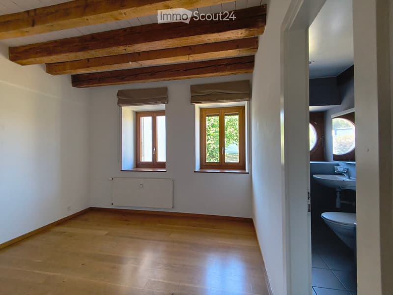 Empty room with wooden flooring, exposed wooden beams on the ceiling, two windows with wooden frames, and a radiator under one window. An adjacent bathroom is partially visible with a sink and toilet.