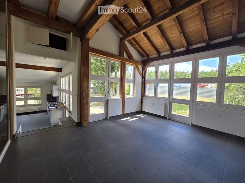 Bright, open room with exposed wooden beams and large windows, featuring a tiled floor and a small kitchen area with a stove and sink.