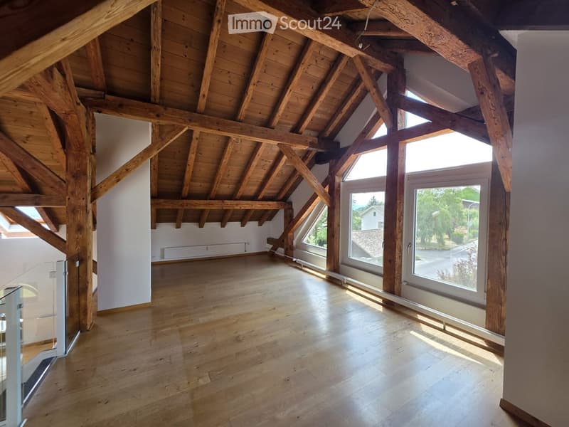 Bright, empty attic space with exposed wooden beams and large windows, featuring wooden flooring and white walls.