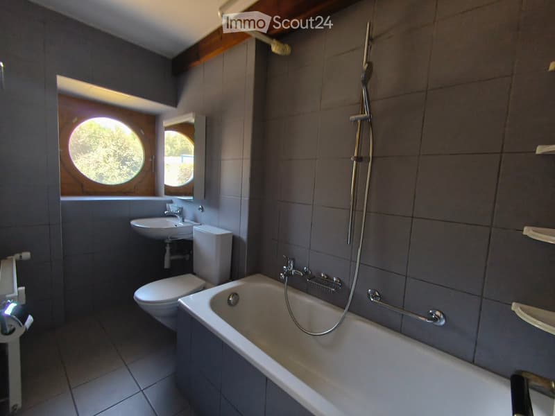 Modern bathroom with a bathtub, toilet, sink, and showerhead. Features a large round window and a built-in shelf.