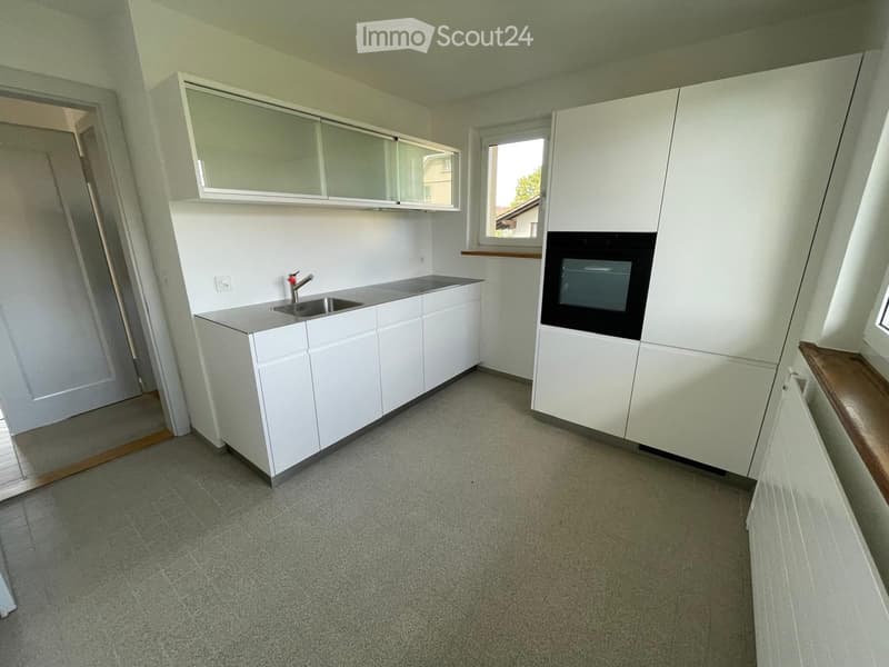 Empty kitchen with white cabinets, a sink, and a window, featuring a grey carpeted floor and a small wooden bench.