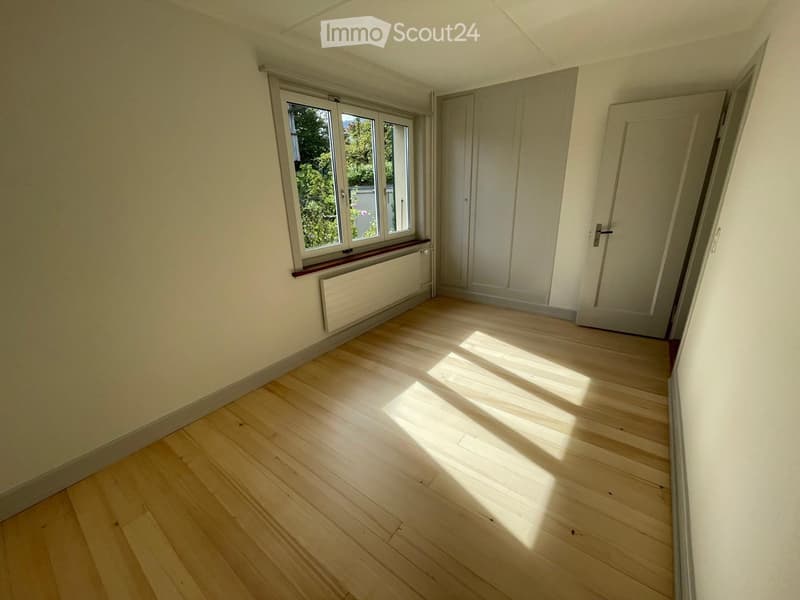 Empty room with wooden flooring, white walls, large window with view of greenery, built-in closet, and radiator under the window.