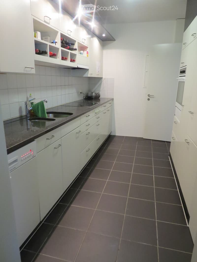 Modern kitchen with white cabinets, dark countertops, and tiled flooring. Features a sink, dishwasher, oven, microwave, and shelves with various items.