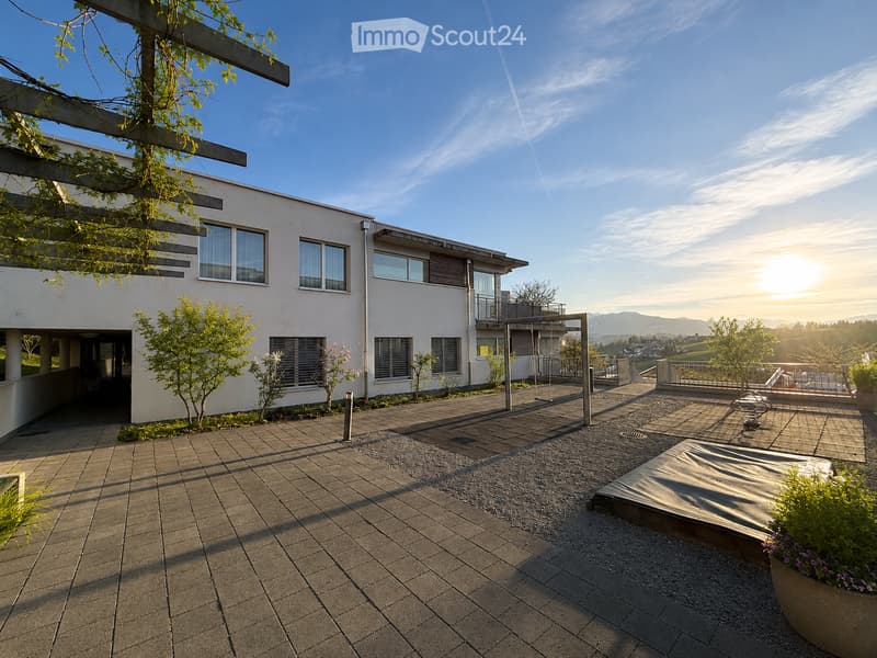 Modern house with a spacious outdoor patio area featuring a wooden deck, gravel section, and potted plants. The house has large windows and a balcony, with a clear blue sky and distant mountains visible.