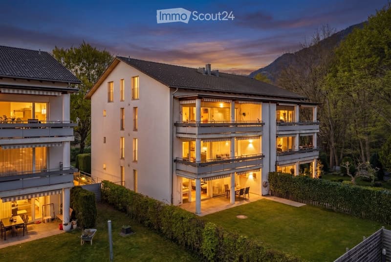 A multi-story residential house with illuminated windows, balconies, and a well-maintained lawn in the foreground.
