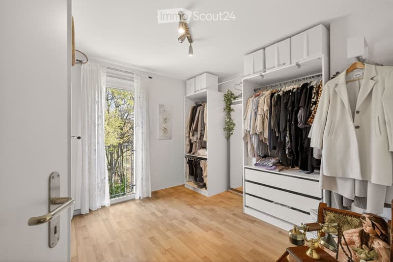 A well-organized walk-in closet with built-in wardrobes, drawers, and shelves. The space features wooden flooring, white walls, and a large window with white curtains. A small table with a vintage telephone and a doll is placed near the door.