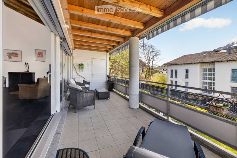 Balcony with wooden ceiling, tiled floor, outdoor furniture including a sofa, chairs, and a small table, with a scenic mountain view.