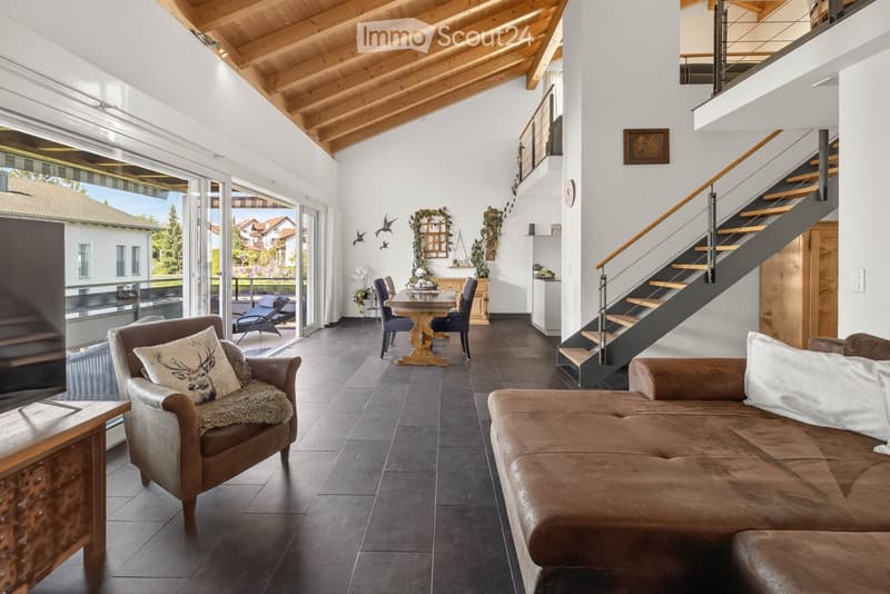 Modern living room with exposed wooden ceiling beams, large windows, and a staircase leading to an upper level. Features include a brown leather sofa, a wooden armchair with a deer pillow, a wooden dining table with chairs, and decorative wall art.