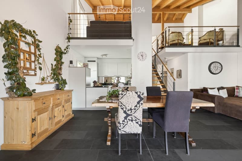 Open-plan living and dining area with a dark tiled floor, wooden dining table, patterned chairs, sofa, and kitchen area with white cabinets and stainless steel appliances.