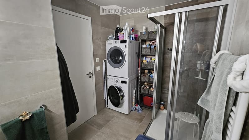 Bathroom with a washing machine, glass shower enclosure, shelves with cleaning supplies, and towels.