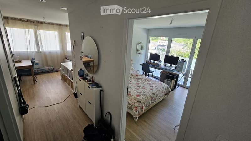A well-lit, modern bedroom with a floral bedspread, a desk with two monitors, a large mirror, and a small dining area with a table and chairs.
