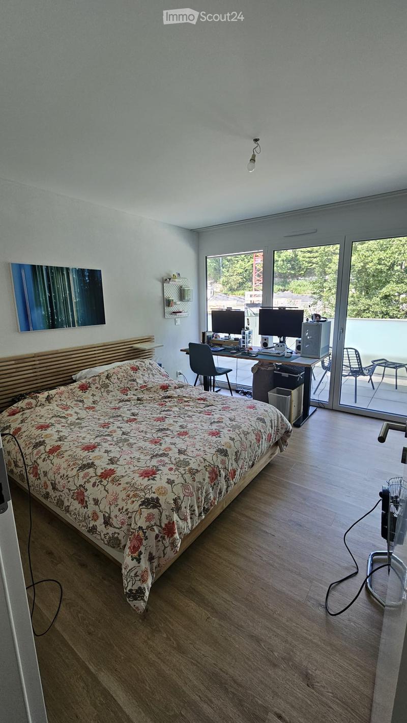 A modern bedroom with a wooden bed, a desk with two monitors, a chair, and a fan. The room has large windows with a view of greenery outside.