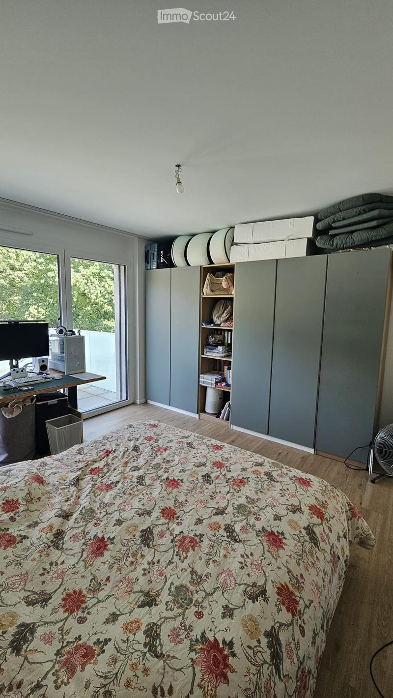 A modern bedroom with a large bed covered in a floral duvet, a wooden desk with a computer setup, and built-in grey cabinets with shelves. Large windows offer a view of greenery outside.