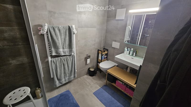 Modern bathroom with grey tiles, a white sink, a mirror, a toilet, a towel rack with towels, a small wooden shelf, and a blue bath mat.