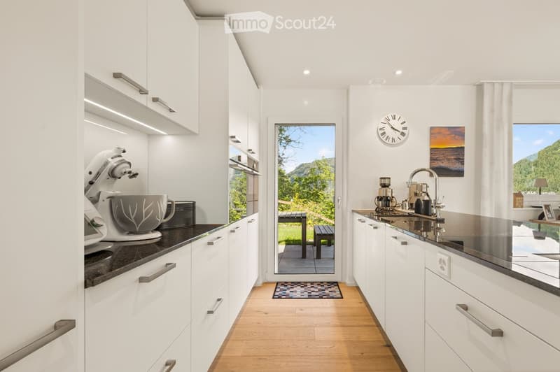 Modern kitchen with white cabinets, black countertops, mixer, coffee maker, and a view of a scenic outdoor area through a large window.