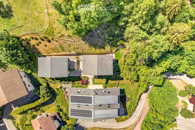 Aerial view of a modern house with solar panels on the roof, surrounded by lush greenery and trees. The house features multiple sections and is situated on a raised ground floor with a driveway leading to it.