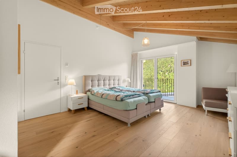 Bright bedroom with wooden flooring, a large bed with a tufted headboard, nightstand with lamp, and a small sofa near a sliding glass door leading to a balcony.