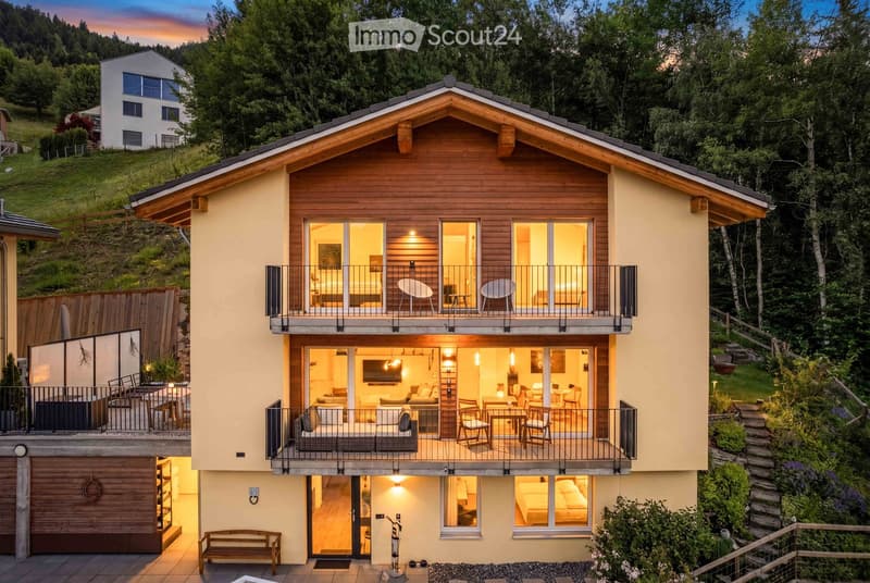 Modern two-story house with wooden accents, large windows, balconies, and a garage. The house is surrounded by greenery and trees.