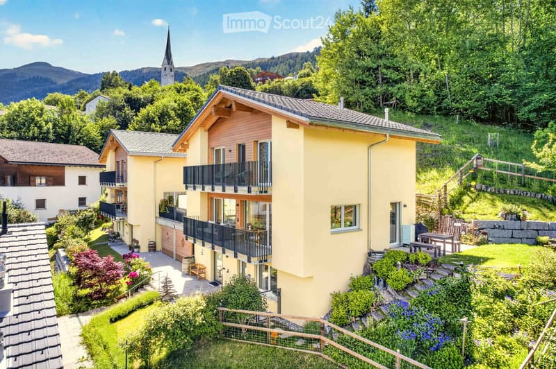 A multi-story yellow house with wooden accents, balconies, and a well-maintained garden in a scenic, green, mountainous area.