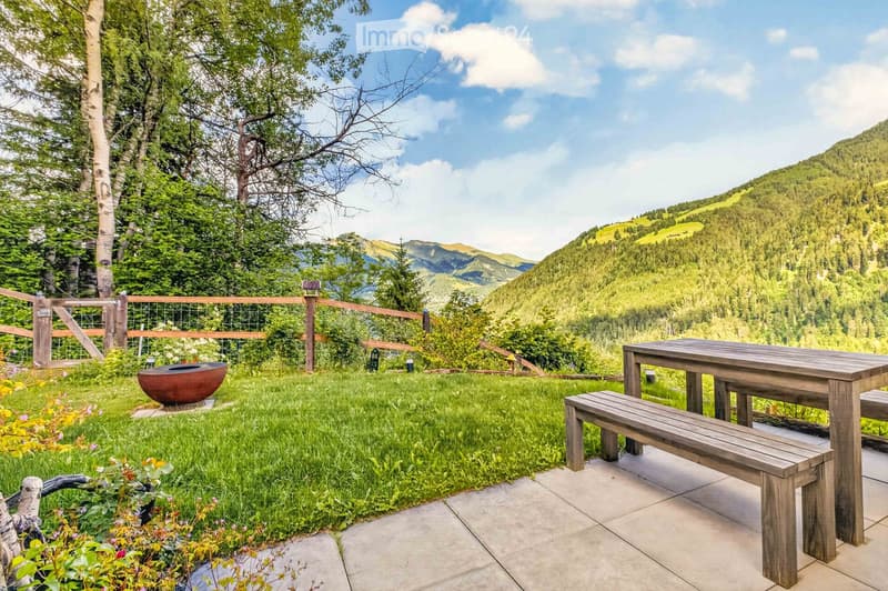 A serene garden area with a wooden table and bench, surrounded by lush greenery and a scenic mountain view.