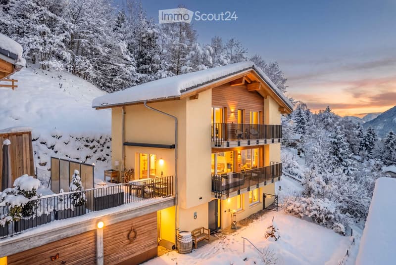 A modern, two-story house with a snow-covered roof and surrounding snowy landscape. The house features large windows, balconies, and a well-lit interior visible through the glass. The exterior includes a wooden garage and a small outdoor seating area.