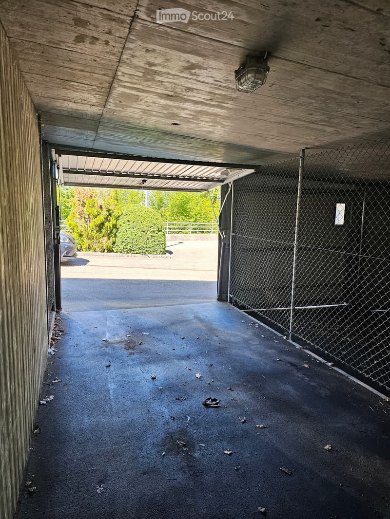 A covered garage area with a concrete floor, metal chain-link fence on one side, and a partially open garage door leading to an outdoor area with greenery visible.