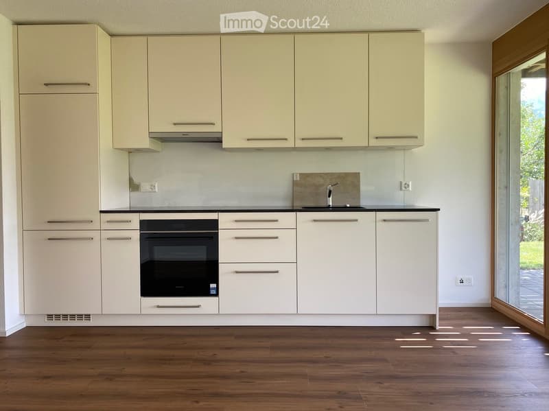 Modern kitchen with light-colored cabinets, stainless steel oven, sink, and faucet, featuring wooden flooring and large windows.