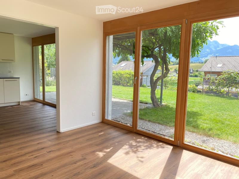 Bright room with wooden flooring, large sliding glass doors opening to a garden, and a small white cabinet visible in the background.