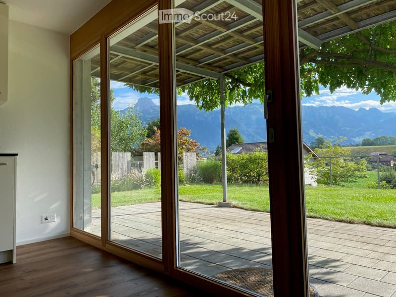 Open sliding glass doors lead to a wooden deck with a view of a green lawn, trees, and distant mountains.