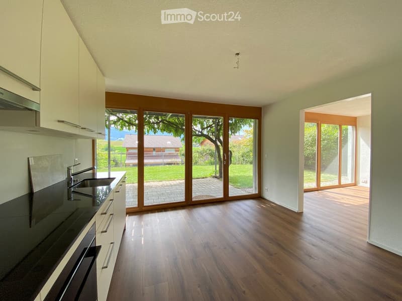 Modern kitchen with light-colored cabinets, dark countertops, and a wooden floor. Large sliding glass doors open to a garden area.