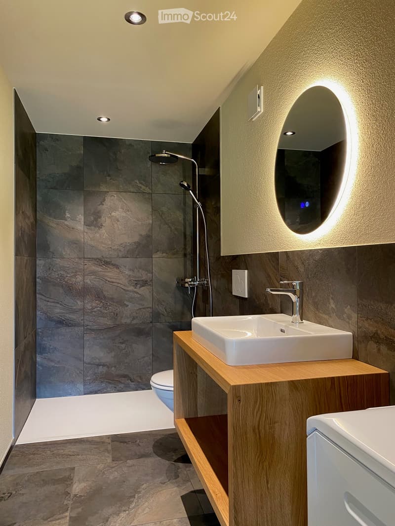 Modern bathroom with a sleek sink, faucet, oval mirror, shower, and toilet. Features marble tiles and recessed lighting.