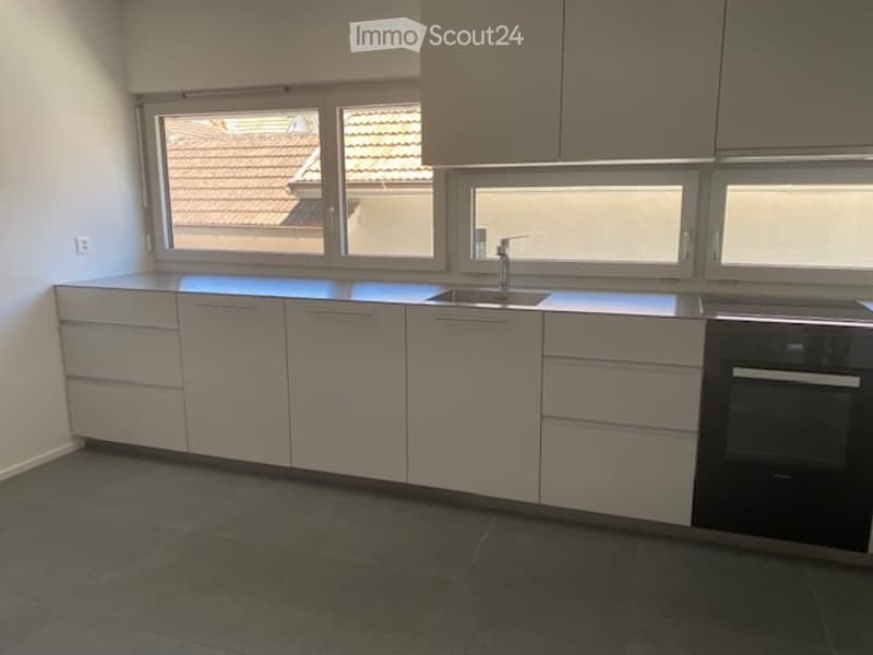 Modern kitchen with white cabinets, dark countertops, and a sink with a faucet. The space features large windows offering a view of a tiled roof.