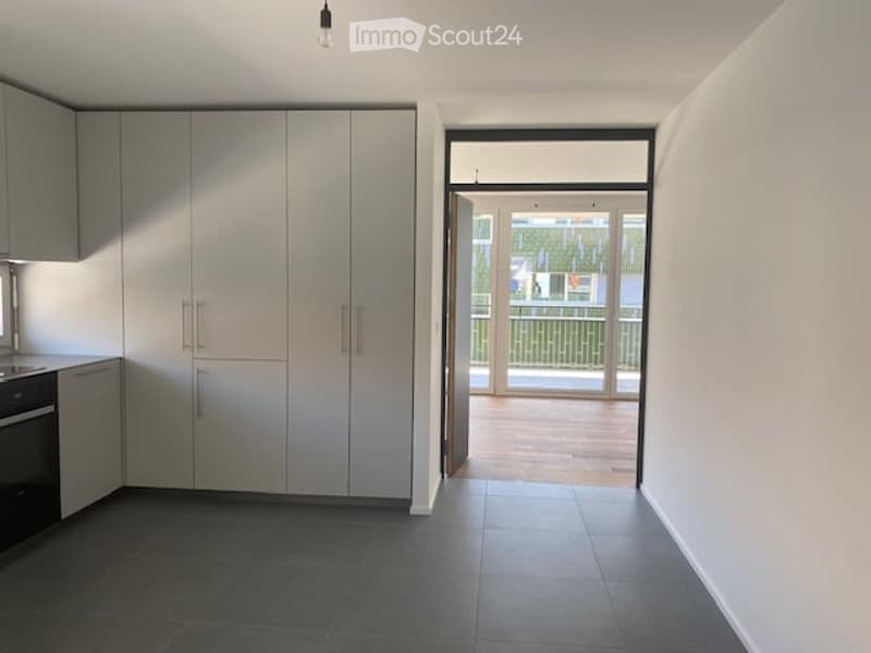Modern kitchen with grey cabinets, dark tiled floor, and an open doorway leading to a balcony with wooden decking.