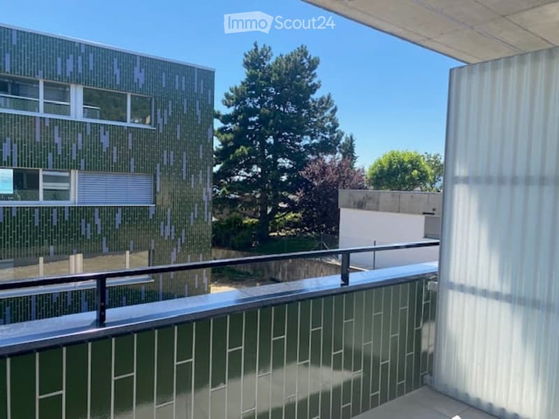 Balcony with a modern railing, overlooking a green area with trees and a contemporary building in the background.