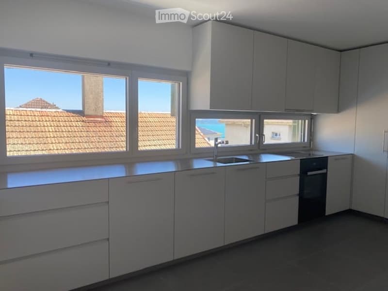 Modern, minimalist kitchen with white cabinets, a central island, and large windows offering a view of the roof and sea.