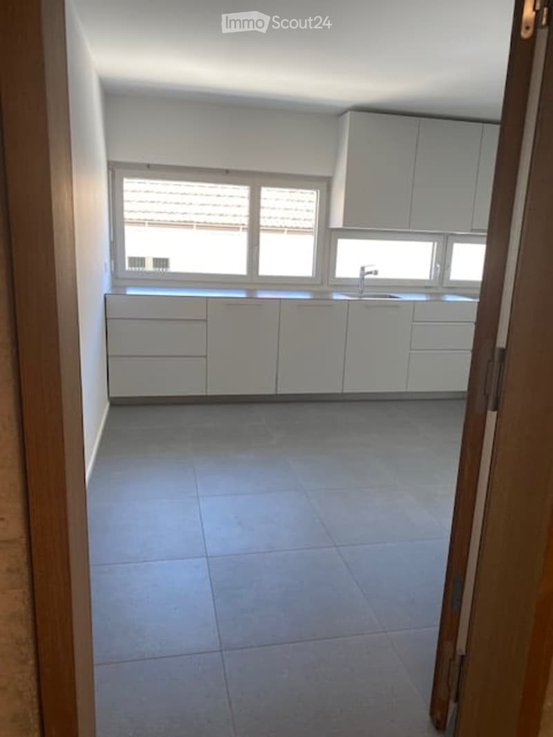 Modern, minimalist kitchen with white cabinets, a central island, and a large window providing natural light. The floor is tiled in a light grey color.