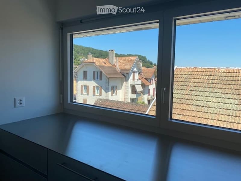 Empty room with large windows offering a view of a town with tiled roofs and surrounding hills. The room has a modern, minimalist design with a grey cabinet and white walls.