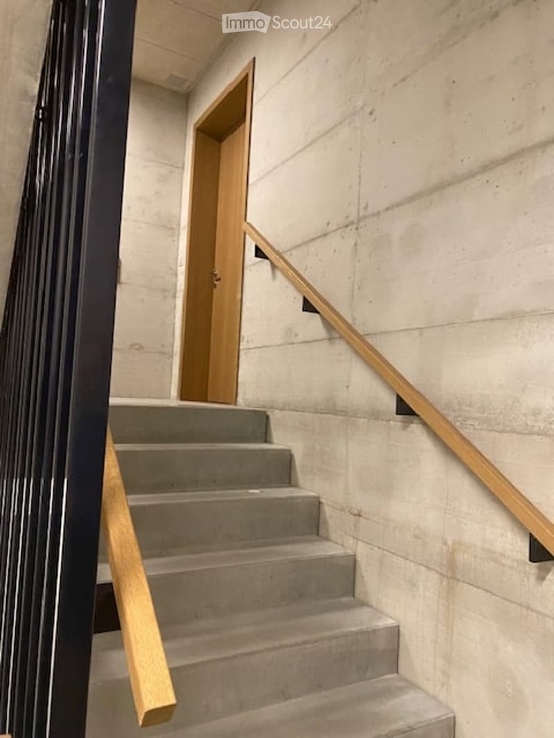 Modern concrete staircase with wooden handrails and a black metal railing, leading to an open wooden door.